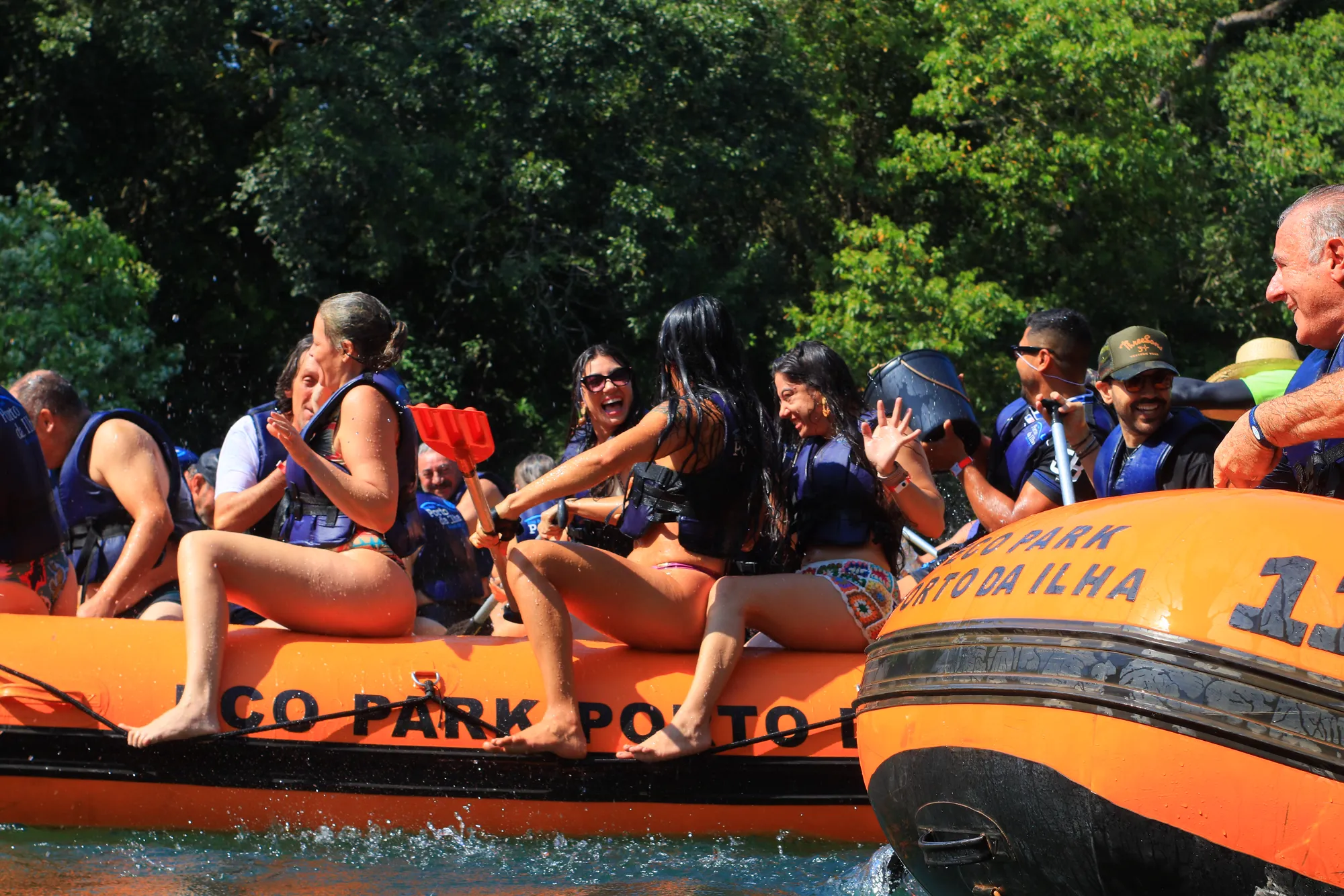 Mulheres sentadas na borda do bote com as pernas para fora, uma acenando, outra com perna estendida, rio e vegetação ao redor