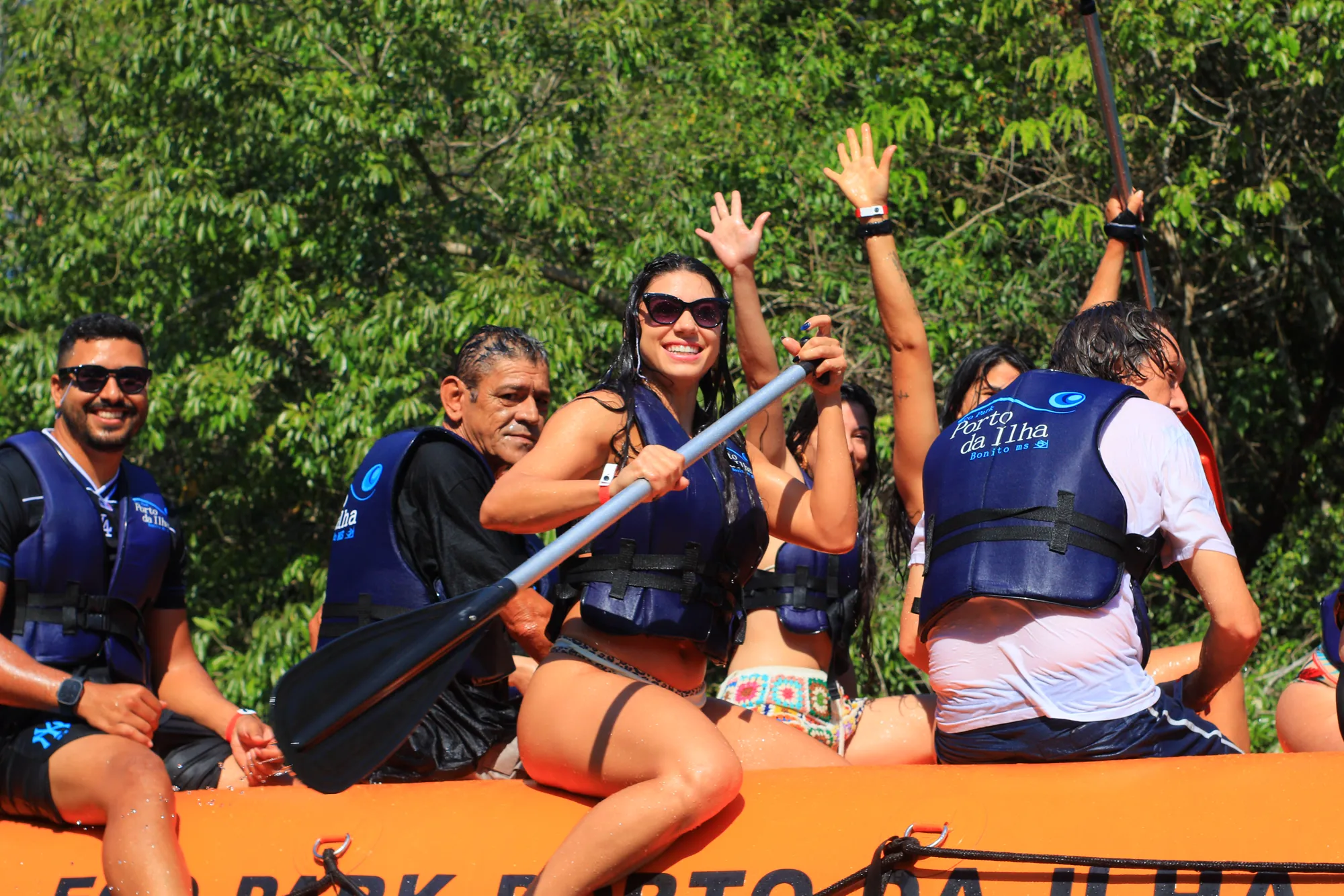 Tripulação do bote de rafting sorrindo: homem de boné virado para trás, mulher apontando o remo para frente, coletes salva-vidas azuis, vegetação densa ao fundo